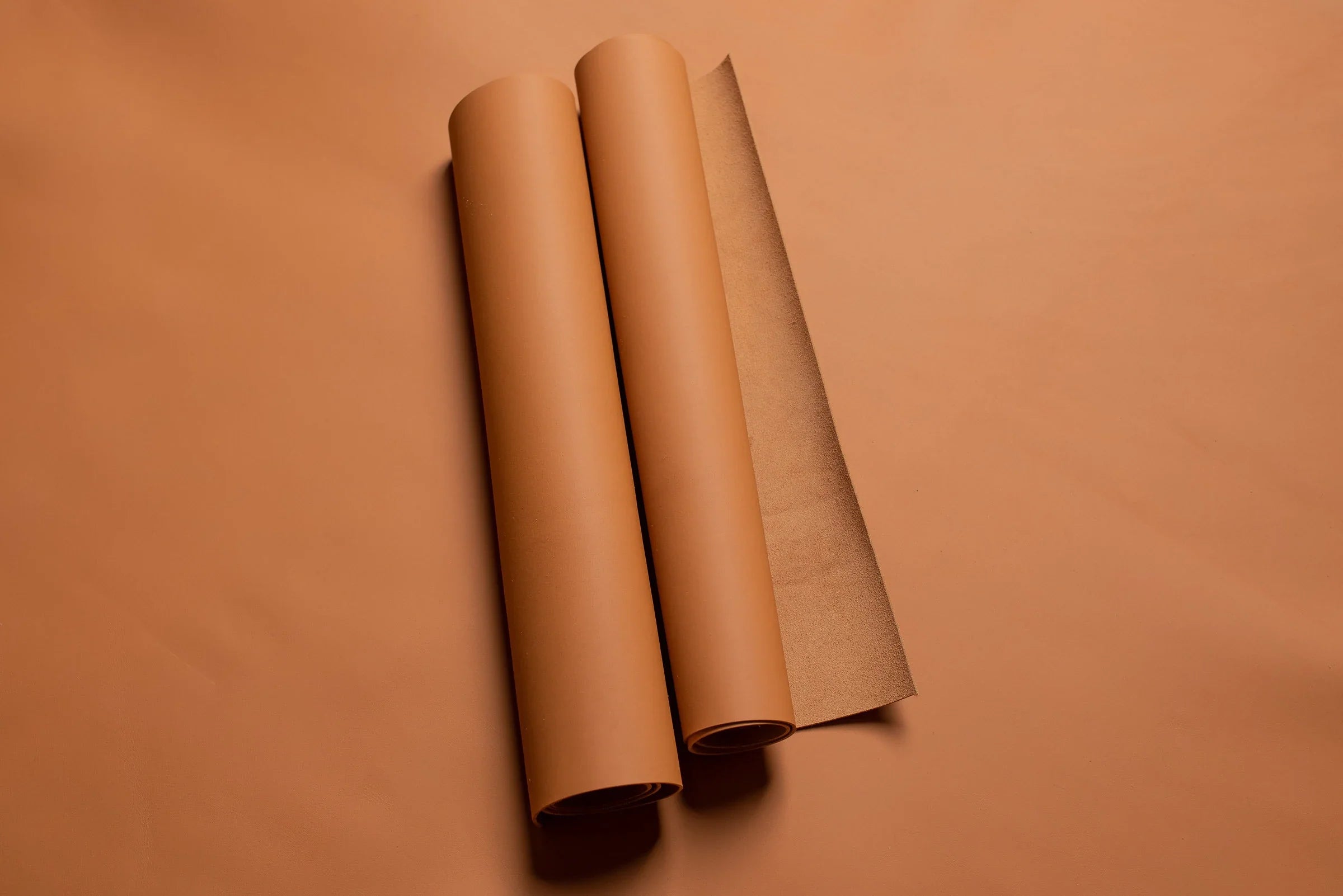 Two full rolls of Barénia leather in a warm caramel hue, laid on a matching leather backdrop, showcasing the smooth surface and natural finish